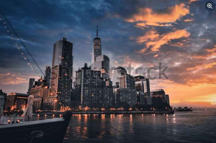 A view of Lower Manhattan’s skyline at sunset, with tall skyscrapers reflecting warm orange and pink hues from the dramatic sky, and calm water in the foreground.